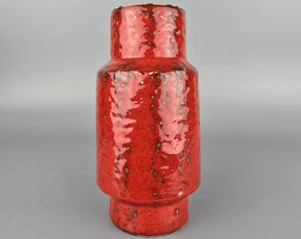 May include: A red ceramic vase with a textured surface. The vase has a cylindrical shape with a wider base and a narrower neck. The glaze has a glossy finish with dark speckles.