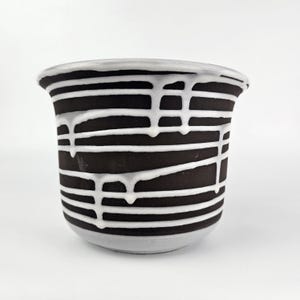 May include: A ceramic pot with a black exterior and horizontal white stripes. White glaze drips down the sides, adding a unique texture. The rim and base are a light grey colour. Suitable for plants or storage, this decorative piece is striking.