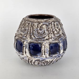 Puede incluir: Un jarrón de cerámica texturizada con forma bulbosa. El jarrón presenta un exterior blanco y azul con detalles cuadrados azul oscuro. La abertura superior es de color marrón oscuro. El jarrón tiene un aspecto único y artesanal.