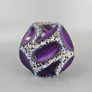 Puede incluir: Un jarrón decorativo de cerámica con un diseño geométrico único. El jarrón presenta un fondo blanco texturizado con motas negras y formas ovaladas y triangulares de color púrpura vibrante. El jarrón tiene un acabado brillante y es una pieza llamativa de decoración del hogar.