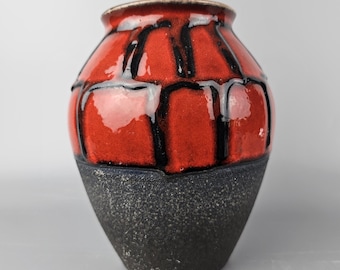 Puede incluir: Un jarrón de cerámica rojo y negro con un diseño geométrico. El jarrón tiene un cuerpo redondeado y una base negra texturizada.