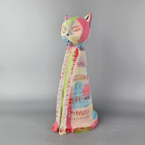 May include: A colourful ceramic cat sculpture with a pink, blue, green and white patterned design. The cat is sitting upright with its head tilted down.