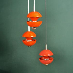 Op de afbeelding: Drie oranje hanglampen met een modern design uit het midden van de eeuw. Elke lamp heeft een afgeronde bovenkant en een komvormige bodem, opgehangen aan witte snoeren. De lampen staan tegen een donkergroene achtergrond.