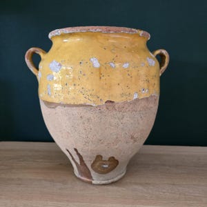 Op de afbeelding: Een vintage keramische pot met twee handvatten, met een geel geglazuurde bovenkant en een bruinbeige basis. De gele glazuur vertoont tekenen van slijtage, met zichtbare afbrokkeling en verkleuring. De pot heeft een afgeronde vorm en een licht uitlopende rand.