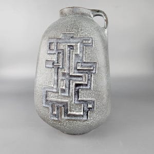 May include: A grey ceramic vase with a textured surface and a geometric design. The vase has a handle and a dark brown rim. The design is a series of interlocking shapes in a darker grey colour.