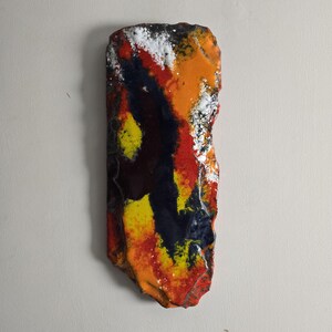 May include: Abstract wall art piece with a rough, irregular shape. The artwork features a vibrant mix of colors, including orange, red, yellow, blue, and white, creating a dynamic and textured appearance. The piece is mounted on a neutral-colored wall.
