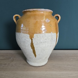 Può includere: Un grande vaso antico in ceramica con due manici. La metà superiore è di un colore giallo dorato caldo, mentre la metà inferiore è di un bianco sporco strutturato. La glassa è colata lungo i lati, creando un aspetto unico e invecchiato. Questo vaso decorativo è adatto per l'arredamento della casa.