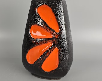 May include: A black ceramic vase with a textured finish, featuring a bold orange floral pattern. The vase has a distinctive form, broader at the top and narrowing towards the bottom. The orange design is glossy, contrasting with the matte black surface.