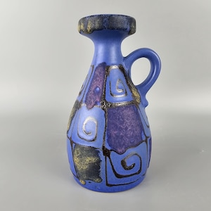 May include: A blue ceramic vase with a geometric pattern in shades of purple, gold, and black. The vase has a handle and a wide mouth.