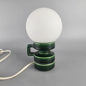 Może przedstawiać: Lampka nocna w stylu vintage z białym kloszem w kształcie kuli i ciemnozieloną ceramiczną podstawą. Podstawa ma uchwyt i warstwową konstrukcję. Lampa jest podłączona białym przewodem.
