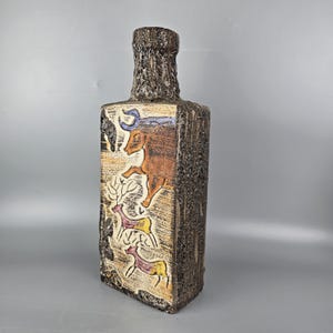 May include: A rectangular, textured brown vase with a narrow neck. The vase features colourful relief carvings of animals, including deer and a bull. The neck has a rough, bark-like texture, and the overall design is rustic and artistic.