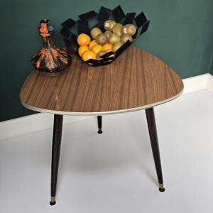 Puede incluir: Una mesa triangular de madera con patas negras y un borde blanco. La mesa tiene un patrón de veta de madera marrón y está coronada con un cuenco de metal negro lleno de kiwis y naranjas.