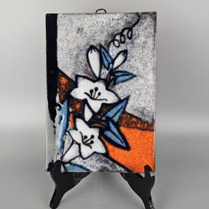 May include: A ceramic wall plaque with a white, blue, and orange floral design. The plaque is mounted on a black easel.