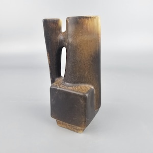 May include: A brown and tan ceramic vase with a unique, geometric design. The vase has a rectangular base, a central body with a handle-like opening, and a tall, vertical section. The color gradient transitions from dark brown to tan.