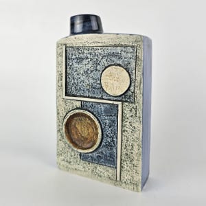 May include: A rectangular ceramic vase with a textured surface. The vase features a dark blue top and a design of squares and circles in shades of blue, beige, and brown. The vase has a modern, geometric aesthetic.