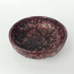 Peut inclure: Un petit bol rond en céramique avec un glaçage moucheté brun foncé et rose. Le bol a un bord légèrement irrégulier et une surface texturée.