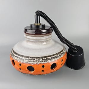 May include: A white and orange ceramic pendant light with a black cord and a black ceiling mount. The light has a textured surface and a ring of holes around the bottom.