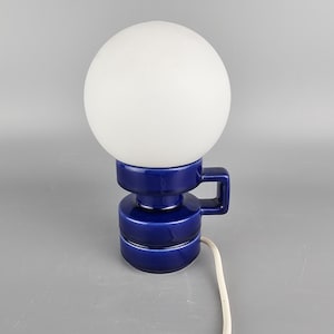 Może przedstawiać: Wintage lampa stołowa z kulistym, białym szklanym kloszem. Podstawa jest z ciemnoniebieskiej ceramiki z uchwytem. Lampa ma biały przewód zasilający.