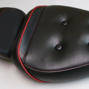 Peut inclure: Selle de moto noire avec surpiqûres rouges et capitonnage. La selle a une texture lisse semblable au cuir et une bordure rouge contrastante. La conception comporte deux zones d'assise séparées.
