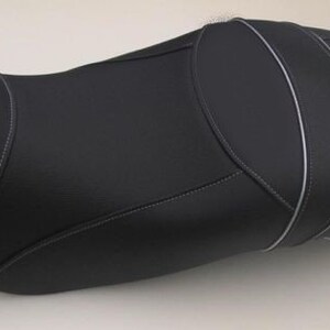 May include: A black motorcycle seat with white stitching and a smooth, textured surface. The seat has a contoured design with a raised back and a rounded front, suitable for a motorcycle.