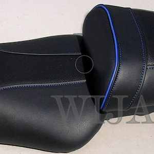 May include: Black motorcycle seat with blue stitching and trim. The seat has a textured surface and a separate passenger section. The design includes a blue accent line around the edges.