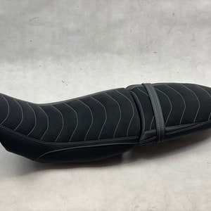 Può includere: Sella da moto nera con cuciture bianche. Il sedile ha un design curvo e una cinghia. Le cuciture formano un motivo ondulato. Il sedile è progettato per il comfort e lo stile, lungo circa 70 cm.