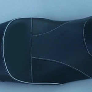 Könnte beinhalten: Ein schwarzer Motorradsitz mit weißen Nähten. Der Sitz hat ein konturiertes Design mit einem erhöhten Mittelteil und einem spitz zulaufenden hinteren Bereich. Die Nähte umreißen die Paneele und Kanten und erzeugen ein kontrastierendes visuelles Detail.