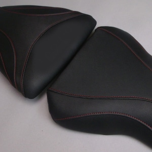 May include: Two black motorcycle seat covers with red stitching. The covers are made of a textured material and have a contoured shape. The stitching follows the edges of the seat covers.