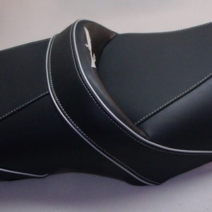 Könnte beinhalten: Schwarzer Motorradsitz mit weißen Nähten und einem silbernen Emblem. Der Sitz hat ein konturiertes Design für Fahrer- und Beifahrerkomfort. Der Sitz besteht aus einem glatten, strapazierfähigen Material.