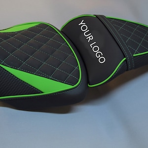 Può includere: Sella da moto nera e verde con cuciture a diamante e superficie strutturata. La sella ha un bordo verde e il testo "YOUR LOGO" in bianco. Progettata per comfort e stile.