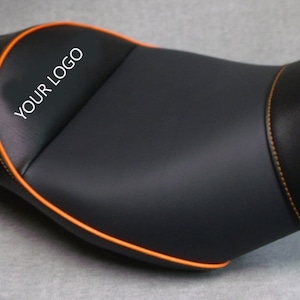 Op de afbeelding: Zwart motorzadel met oranje stiksels en afwerking. De zitting heeft een glad, gewatteerd oppervlak met een voorgevormd ontwerp. De tekst "YOUR LOGO" is op de zitting gedrukt.