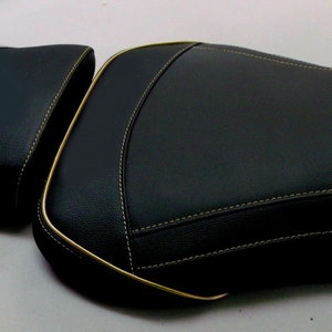 May include: Two-piece black motorcycle seat with gold trim and white stitching. The seat has a textured surface and a contoured design for rider comfort. The seat is designed for a motorcycle.