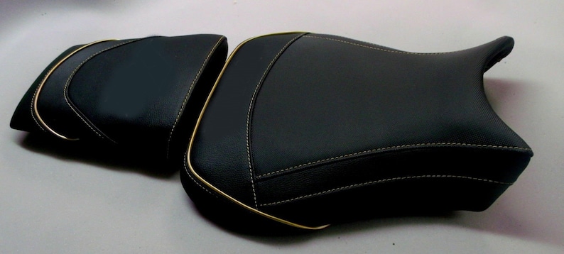 May include: Two-piece black motorcycle seat with gold trim and white stitching. The seat has a textured surface and a contoured design for rider comfort. The seat is designed for a motorcycle.