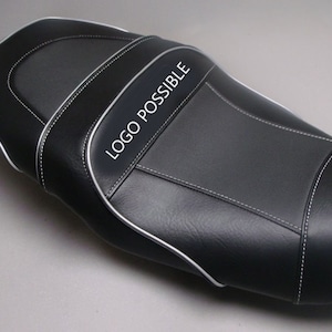 May include: A black motorcycle seat with white stitching and a silver trim. The seat has a textured surface and the text "LOGO POSSIBLE" is printed on the side. The seat is designed for comfort and style.