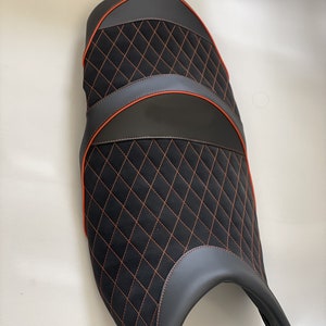 May include: A motorcycle seat featuring a black, diamond-stitched central panel and dark grey side panels. It has orange stitching and trim, and a black base. The seat is designed for comfort and style.