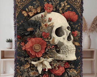 May include: A decorative throw blanket featuring a skull surrounded by red, black, and white flowers. The design includes butterflies and ornate floral patterns on a dark background. The blanket has a fringed edge.