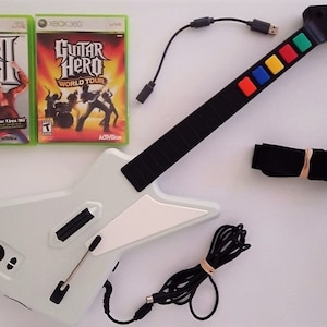 May include: A white and black Gibson guitar controller for Xbox 360, with colored buttons and a black strap. Two Guitar Hero game cases are also included.