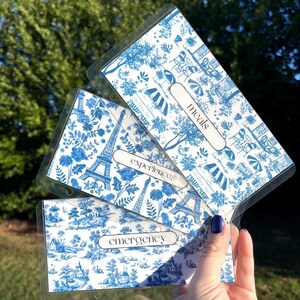 K&ouml;nnte beinhalten: Drei transparente Plastik-Taschen mit blauen und wei&szlig;en Toile-Muster-Eins&auml;tzen. Die Taschen haben L&ouml;cher f&uuml;r einen Ordner. Die Taschen sind mit "Mahlzeiten", "Erlebnisse" und "Notfall" beschriftet.