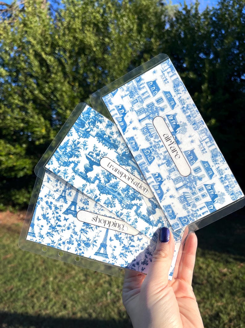 K&ouml;nnte beinhalten: Vier blaue und wei&szlig;e gemusterte Budget-Umschl&auml;ge mit durchsichtigen Plastikh&uuml;llen. Die Umschl&auml;ge sind mit "Flugkosten", "Transport", "Einkaufen" und "Sonstiges" beschriftet.