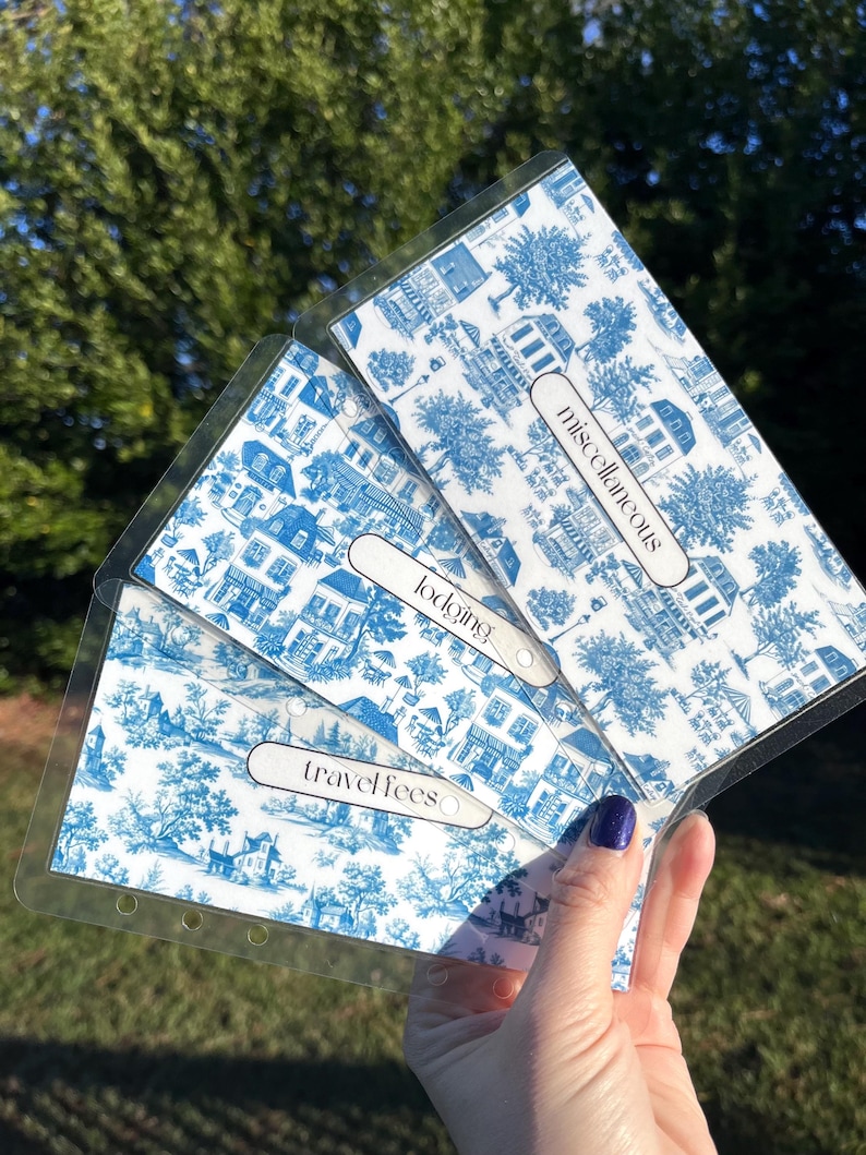 K&ouml;nnte beinhalten: Drei transparente Plastik-Seitensch&uuml;tzer mit blauen und wei&szlig;en Toile-Muster-Papiereins&auml;tzen. Die Eins&auml;tze sind mit "Reisekosten", "Unterkunft" und "Sonstiges" beschriftet.
