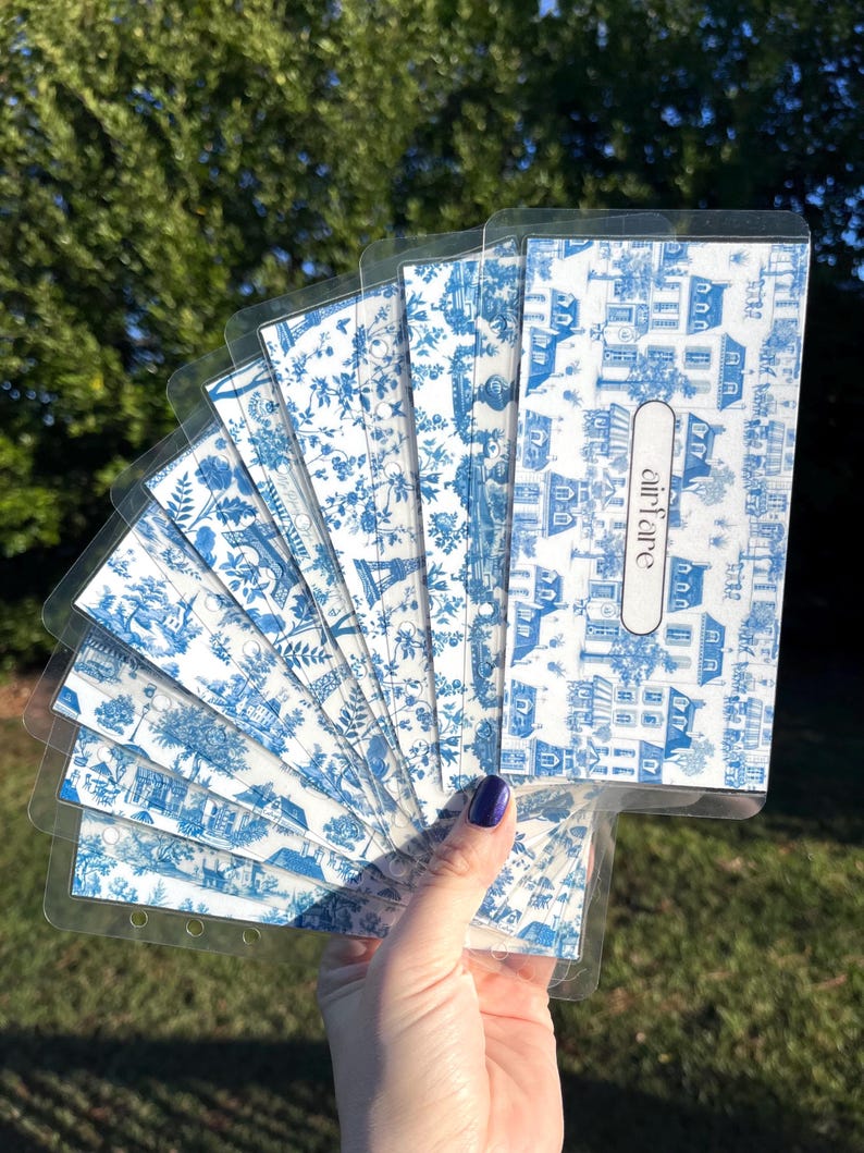 K&ouml;nnte beinhalten: Ein Satz von acht blauen und wei&szlig;en gemusterten Planer-Teilern mit durchsichtigen Plastikh&uuml;llen. Die Teiler zeigen ein sich wiederholendes Muster aus H&auml;usern und Blumen. Das Wort "airfare" ist auf einem der Teiler gedruckt.