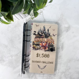 Op de afbeelding: Een doorzichtige binder met een zwarte rug bevat een beige spaaruitdaging-inzet. De inzet toont een Universal Studios-afbeelding, de tekst "$1.500 Savings Challenge" en een gevleugeld embleem. Groene bladeren zijn zichtbaar op de achtergrond.