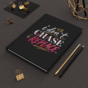 May include: A black hardcover notebook featuring the white and pink text "i don't CHASE i REPLACE" with a small gold crown and heart graphic. The notebook is positioned next to two pencils and several gold-toned metal binder clips.