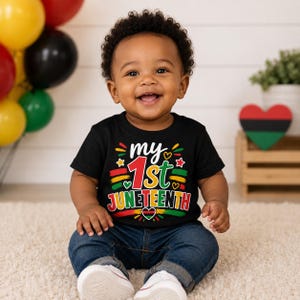 May include: A black short-sleeved t-shirt featuring the text "my 1st JUNETEENTH" in red, yellow, and green lettering. The design includes small star and heart icons in the same colors, with a heart containing a red, black, and green flag.