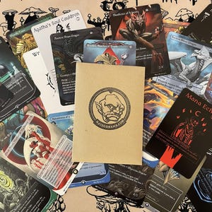May include: A collection of fantasy trading cards featuring various illustrations and text, including "Agatha's Soul Cauldron" and "Mana Echoes." A brown paper envelope with a circular logo and "NATEGODRAWS.COM" is in the center.