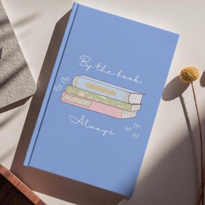May include: A light blue hardback journal featuring a central illustration of a stack of three books in light blue, green, and pink. The text "By the book." appears above the stack, and "Always." appears below, accented by small heart outlines.