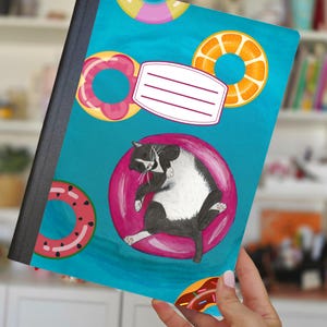 May include: A notebook with a turquoise cover featuring a black and white cat lounging on a pink inflatable ring. The cover also displays various colorful donut-shaped floaties. A white label with lines is present for personalization.