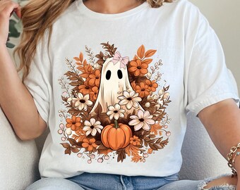May include: White t-shirt featuring a cartoon ghost with a floral arrangement of orange, brown, and white flowers, and a small pumpkin. The ghost has a pink bow. The shirt is a casual, comfortable style.