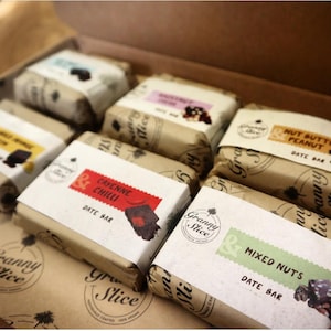 May include: A selection of date bars in a wooden box, each individually wrapped in brown paper. Flavours include "Cayenne Chilli" and "Mixed Nuts", with the "Granny Slice" logo on the packaging. The bars are presented in a rustic style.