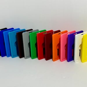 May include: A set of 14 plastic badge holders in various colors, including black, blue, teal, maroon, gray, green, red, orange, pink, purple, white, and yellow. Each holder has a rectangular shape with a cut-out for a badge.